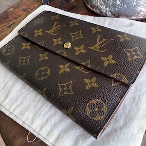 May include: Brown wallet with a flap closure. The wallet features a repeating pattern of interlocking logos and floral designs in a lighter brown color. A gold-colored snap secures the flap. The wallet is resting on a white cloth.