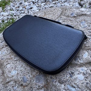 May include: A black, rectangular zippered case with a textured surface. The case is closed and appears to be made of leather or a similar material. The zipper is black and runs around the perimeter of the case.