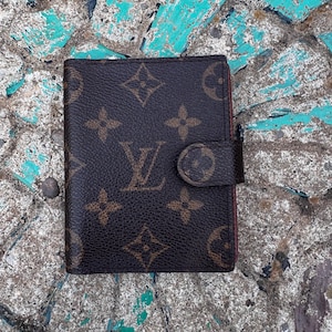 May include: A brown leather wallet featuring the iconic Louis Vuitton monogram pattern in a dark brown and tan color scheme. The wallet has a rectangular shape with a secure snap-button closure tab on the right side, showcasing a textured finish.