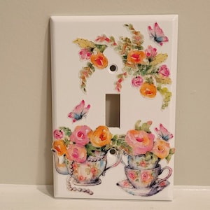 May include: White light switch cover with a floral design featuring pink, yellow, and blue flowers, butterflies, and two teacups with floral patterns.