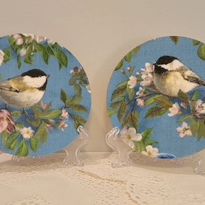 May include: Two round ceramic plates with a blue background and a white bird perched on a branch with pink and white flowers. The plates are on clear plastic stands.