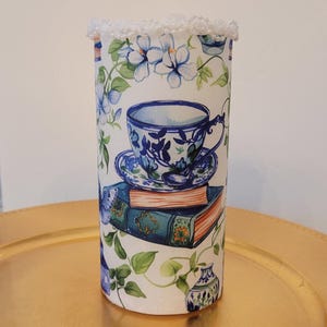May include: White ceramic cylinder vase with a blue and white floral pattern. The vase features a teacup and saucer with a floral design, a stack of books, and a small vase with a floral design. The vase has a white lace trim around the top.