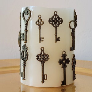 May include: A white candle with various antique-style key charms attached to it. The keys are made of metal and have a bronze finish.