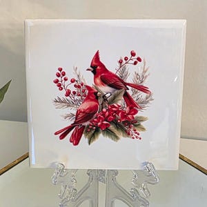 May include: A white ceramic tile with a red cardinal bird and holly berry design. The tile is on a white stand.