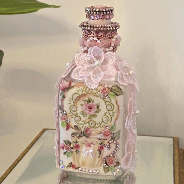 May include: A decorative bottle with a floral design. The bottle is adorned with pink lace, pearls, and rhinestones. The stopper is embellished with pink rhinestones. The bottle features a painted floral motif with roses and a decorative frame.