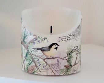 May include: A white candle with a bird and pine branch design. The candle has a black wick and is in a curved shape. The text on the candle reads "In the air a little black cat! and a bell!"