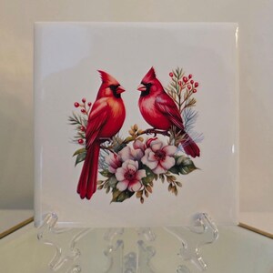 May include: A white ceramic tile with a painted image of two red cardinals perched on a branch with white flowers. The cardinals are facing each other and have red beaks and black eyes.