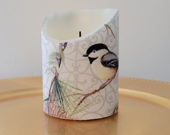 May include: A white pillar candle with a design of a bird on a branch with pine needles and berries. The design is on a light gray background with a white and gray floral pattern.