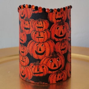 May include: A black cylindrical candle holder with a Halloween theme. The holder is covered in a fabric with a repeating pattern of orange jack-o'-lantern faces. The top edge of the holder is decorated with a row of orange and black rhinestones.
