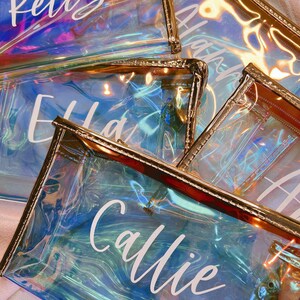 May include: Four iridescent clear plastic zippered pouches with gold trim. Each pouch has a white name written on it: Kelly, Adam, Ella, and Callie.