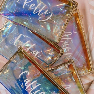 May include: Four iridescent clear zippered pouches with gold trim. Each pouch has a name written on it in white: Kelly, Ella, Callie, and Anna.