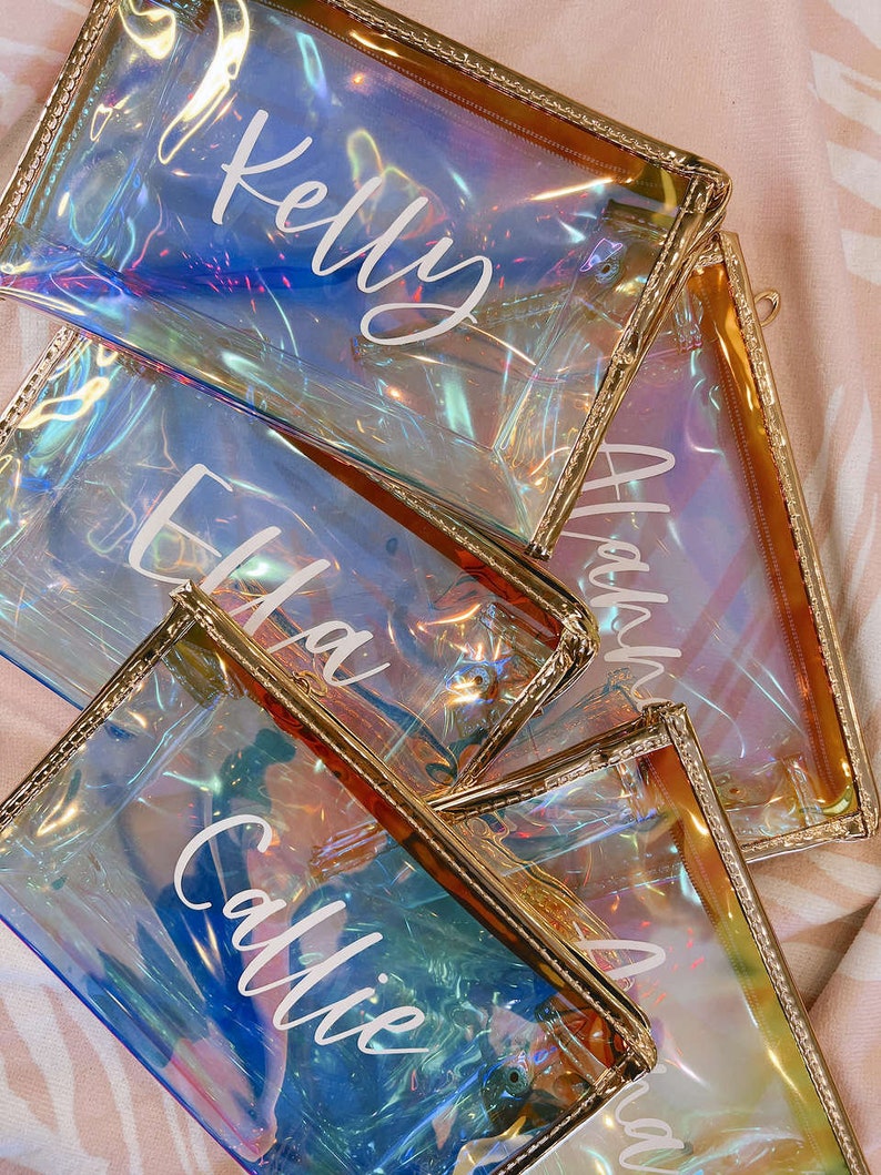 May include: Four iridescent clear zippered pouches with gold trim. Each pouch has a name written on it in white: Kelly, Ella, Callie, and Anna.
