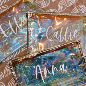 May include: Three iridescent clear plastic zippered pouches with gold trim. The pouches have the names "Ella", "Callie", and "Anna" printed in white on them.