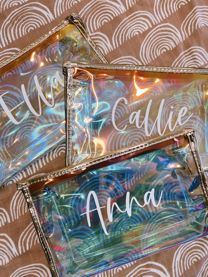 May include: Three iridescent clear plastic zippered pouches with gold trim. The pouches have the names "Ella", "Callie", and "Anna" printed in white on them.