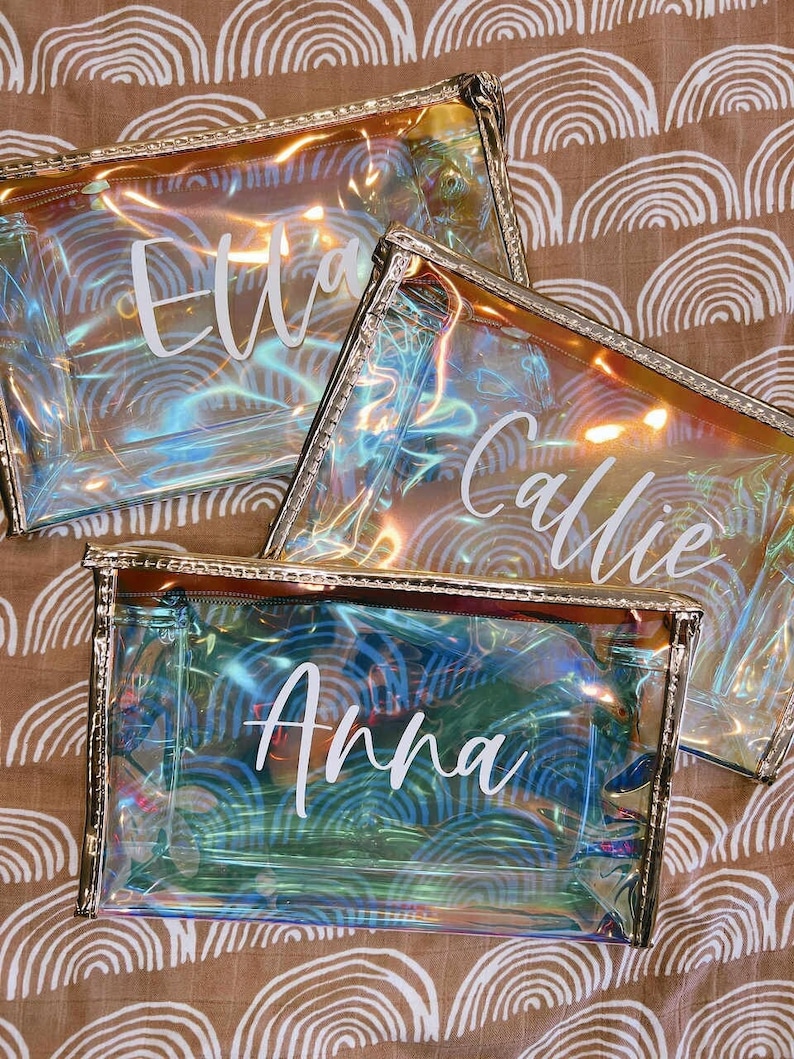 May include: Three iridescent clear zippered pouches with gold trim. The pouches are personalized with the names Ella, Callie, and Anna in white lettering.