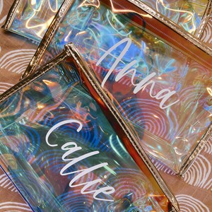 May include: Three iridescent clear plastic zippered pouches with gold trim. The pouches have the names "Anna", "Callie" and a third name that is not visible in the image, printed in white on the front.