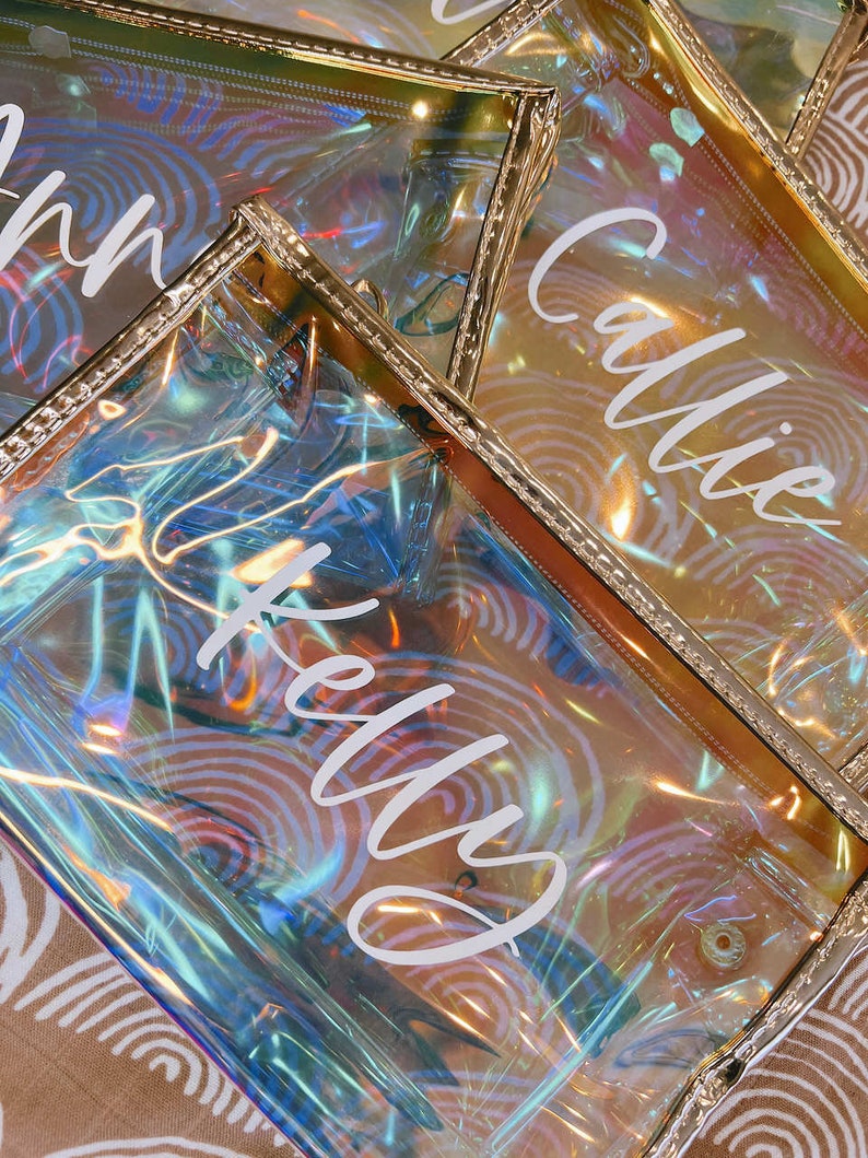 May include: Three iridescent clear plastic zippered pouches with gold trim. The pouches have the names "Kelly", "Callie", and "Ann" written in white on them.