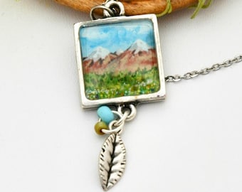May include: Silver-toned necklace featuring a square pendant with a miniature landscape painting of mountains, blue sky, and green fields. A small leaf charm and colorful beads dangle below the pendant. The chain is delicate and the overall design is nature-inspired.