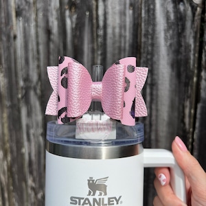 Puede incluir: Un vaso Stanley blanco con un adorno en forma de lazo rosa. El lazo tiene un estampado de leopardo negro. El vaso tiene un asa y el logotipo de Stanley es visible en la parte delantera.