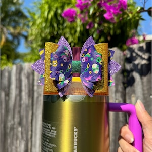 Könnte beinhalten: Ein goldfarbener Edelstahl-Reisebecher mit lila Griff, verziert mit einer dekorativen Schleife. Die Schleife hat Schichten aus goldenem Glitzer, lila Glitzer und einem gemusterten Stoff mit Mardi-Gras-Maskenmotiven. Der Becher wird von einer Person gehalten.