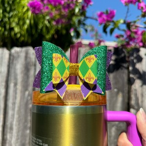 Könnte beinhalten: Ein goldener Edelstahl-Reisebecher mit lila Griff und einer dekorativen Schleife. Die Schleife hat grüne Glitzerflügel, eine gelb-grüne Mitte mit einem Fleur-de-Lis-Muster und lila Akzente. Der Becher hat einen Strohhalm.