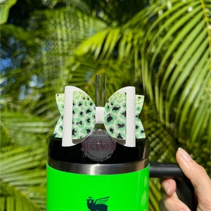 Puede incluir: Un vaso Stanley verde brillante con un asa negra y el logotipo de Stanley. Un lazo decorativo con estampado de leopardo blanco y verde está sujeto a la parte superior. Se inserta una pajita transparente en el vaso.
