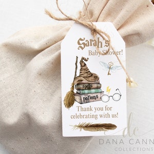 May include: A white favor tag with a brown and gold illustration of a witch's hat, a broom, and books with the words "Spells" and "Potions" on them. The text "Sarah's Baby Shower!" is at the top of the tag and "Thank you for celebrating with us!" is at the bottom. The tag also has a small illustration of a dragonfly and a key. The text "DANA CANNON COLLECTIONS" is at the bottom of the tag.