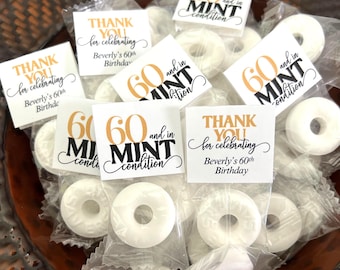 May include: A collection of individually wrapped white ring-shaped mint candies. Each clear plastic wrapper features a square white tag with black and gold text reading either "THANK YOU for celebrating Beverly's 60th Birthday" or "60 and in MINT condition."