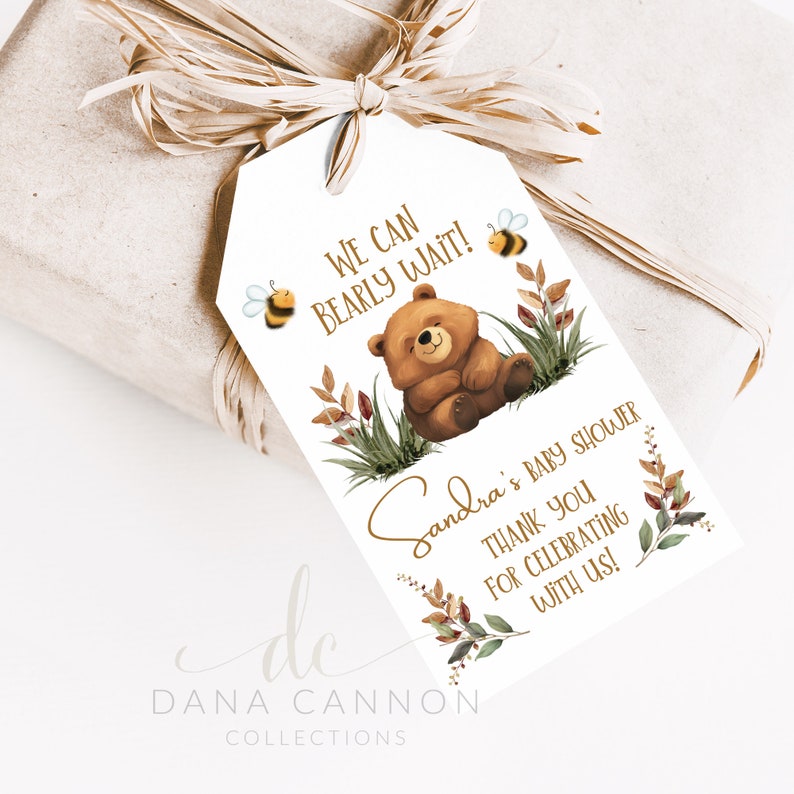 May include: A white gift tag with a brown bear illustration and the text "We can bearly wait!"  The tag also features a floral design with bees and the text "Sandra's Baby Shower Thank you for celebrating with us!"