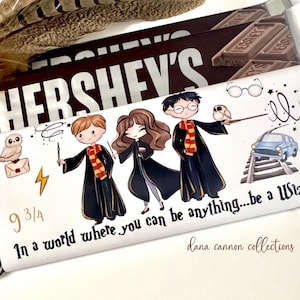 May include: A white chocolate bar wrapper with a Harry Potter theme. The wrapper features a cartoon illustration of three children dressed as witches and wizards, a Hogwarts Express train, and an owl. The text on the wrapper reads "9 3/4 In a world where you can be anything...be a Wizard!" and "dana cannon collections".