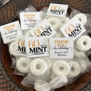 May include: A close-up shot of a collection of white mint candies in clear plastic wrappers. Each wrapper has a small square label with gold and black text that reads "60 and in MINT condition" and "Thank You for celebrating Beverly's 60th Birthday."