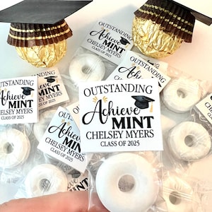 May include: A set of graduation party favors featuring small white mints wrapped in clear cellophane bags. Each bag has a white tag with black text that reads "Outstanding Achieve Mint Chelsey Myers Class of 2025". The tags also feature a small black graduation cap.