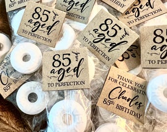 May include: A wooden bowl filled with small clear plastic bags containing white mint candies. Each bag has a small brown tag that reads "85 and aged to perfection" and "Thank you for celebrating Charles 85th Birthday".