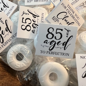 May include: Close-up of individually wrapped white mint candies. Each candy is packaged in a clear plastic wrapper. Small white tags with black text read "Thank you for celebrating Charles 85th Birthday" and "85 and aged to perfection."