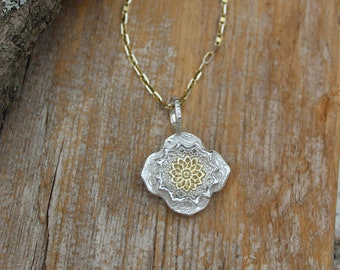 May include: A silver and gold necklace with a detailed mandala pendant. The pendant is a circular design with a smaller gold circle in the centre. The necklace is on a wooden surface with a purple flower.