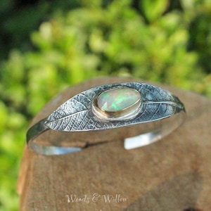 May include: A silver leaf cuff bracelet with a green opal gemstone set in a gold bezel. The bracelet is textured to resemble a leaf.