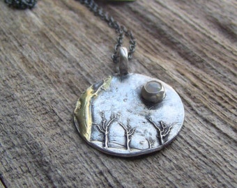 Könnte beinhalten: Eine silberne Halskette mit einem runden Anhänger, der ein strukturiertes Design von Bäumen und einem kleinen Mondstein zeigt. Der Anhänger ist an einer silbernen Kette befestigt.