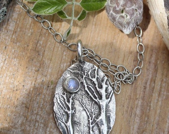 Könnte beinhalten: Eine silberne Halskette mit einem Anhänger, der einen Mondstein und ein strukturiertes Design von Bäumen zeigt. Der Anhänger ist oval geformt und hängt an einer silbernen Kette.