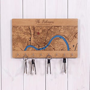 May include: Wooden key holder with a detailed map design, featuring a blue river and city layout. The text "The Robinsons" and coordinates are engraved at the top. Includes several key hooks and keys hanging below. The wood has a natural finish.