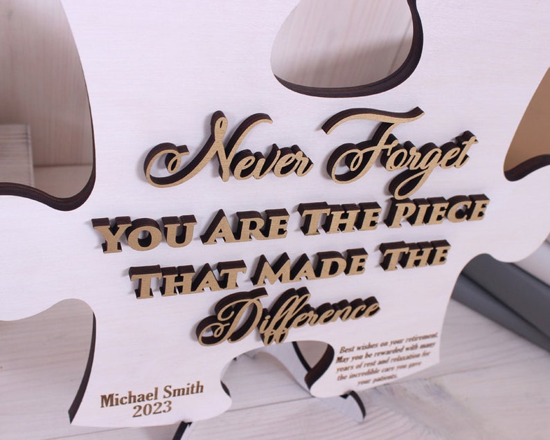 Peut inclure: Pi&egrave;ce de puzzle en bois blanc avec le texte "Never Forget You Are The Piece That Made The Difference" en lettres dor&eacute;es. Le texte "Michael Smith 2023" est grav&eacute; au bas de la pi&egrave;ce.
