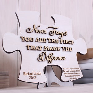 Peut inclure: Une pi&egrave;ce de puzzle en bois blanc avec le texte "Never Forget You Are The Piece That Made The Difference" en lettres dor&eacute;es. Le bas de la pi&egrave;ce porte le texte "Michael Smith 2023" et un message sur la retraite.
