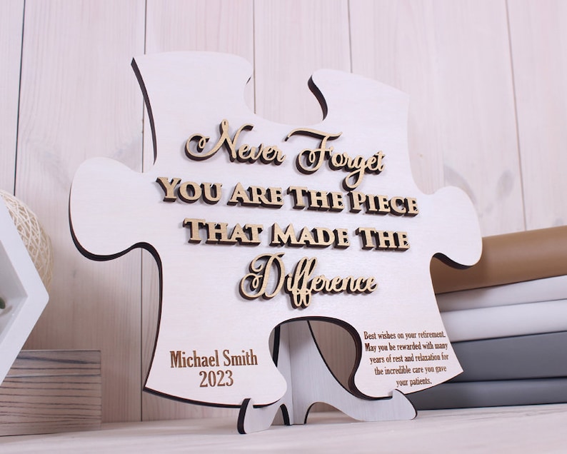 Peut inclure: Une pi&egrave;ce de puzzle en bois blanc avec le texte "Never Forget You Are The Piece That Made The Difference" en lettres dor&eacute;es. Le bas de la pi&egrave;ce porte le texte "Michael Smith 2023" et un message sur la retraite.