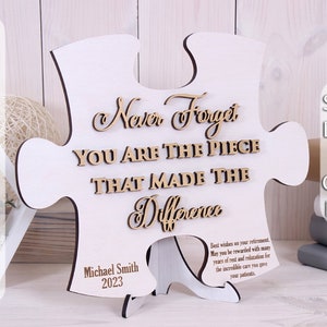 Op de afbeelding: Een wit houten puzzelstuk met de tekst "Never Forget You Are The Piece That Made The Difference" in bruine letters. Het puzzelstuk staat op een kleine houten standaard. De tekst "Michael Smith 2023" is gegraveerd op de onderkant van het puzzelstuk. De tekst "Beste wensen met uw pensioen. Moge u worden beloond met vele jaren rust en ontspanning voor de ongelooflijke zorg die u aan uw patiënten hebt gegeven." is gegraveerd op de zijkant van het puzzelstuk.