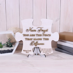 Peut inclure: Une pi&egrave;ce de puzzle en bois blanc en forme de puzzle avec les mots "Never Forget You Are The Piece That Made The Difference" en lettres brunes. Le bas de la pi&egrave;ce porte le nom "Michael Smith" et l'ann&eacute;e "2023" en lettres noires.