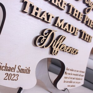 Peut inclure: Une plaque en bois blanc avec le texte "You are the piece that made the difference" en lettres brunes. La plaque est en forme de pi&egrave;ce de puzzle et porte le nom "Michael Smith" et l'ann&eacute;e "2023" grav&eacute;s en bas.
