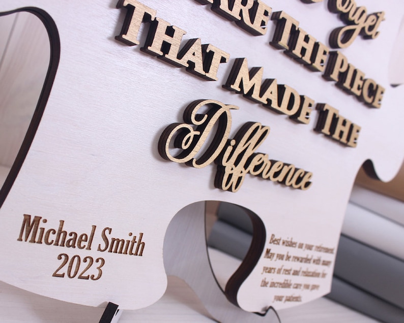 Peut inclure: Une plaque en bois blanc avec le texte "You are the piece that made the difference" en lettres brunes. La plaque est en forme de pi&egrave;ce de puzzle et porte le nom "Michael Smith" et l'ann&eacute;e "2023" grav&eacute;s en bas.