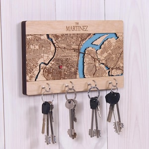 Könnte beinhalten: Ein rechteckiger Wand-Schlüsselhalter aus Holz mit einer lasergeschnittenen Karte von Philadelphia und Camden sowie einer blauen Wassereinlage. Der Text THE MARTINEZ ist oben eingraviert und ein kleines rotes Herz markiert einen Ort. Vier Metallhaken.