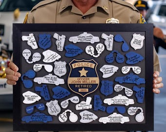 Könnte beinhalten: Ein gerahmter Ruhestands-Schattenkasten mit Polizei-Themen-Ausschnitten. Die Box zeigt ein zentrales Polizeilabzeichen mit dem Namen "Officer John Miller" und "Retired 2025". Um das Abzeichen herum befinden sich blaue und weiße Ausschnitte von Polizeiautos, Handschellen und Sprechblasen.