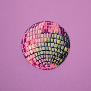 May include: A colorful, embroidered patch in the shape of a disco ball. The patch features a mosaic of small squares in shades of pink, purple, yellow, and green, with a pink border. The background is a solid lavender color.