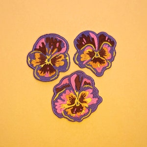 May include: Three embroidered pansy patches in shades of purple, brown, pink, yellow, and orange. The patches are arranged on a yellow background. The patches are likely for clothing or craft projects.